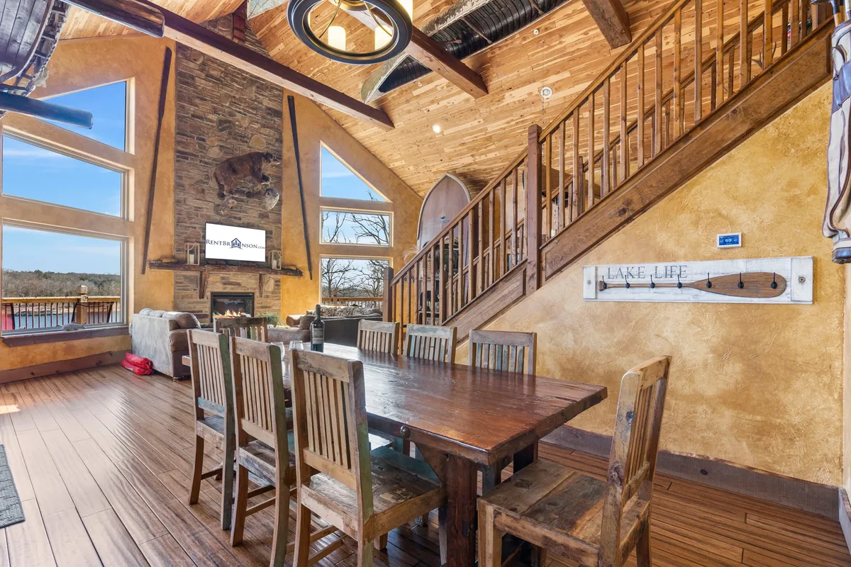 A warmly lit open-concept living and dining area anchored by a large dining table, cozy stone fireplace, and expansive windows that reveal scenic lake views. The space features inviting rustic decor and wooden details, perfect for group gatherings and shared meals.