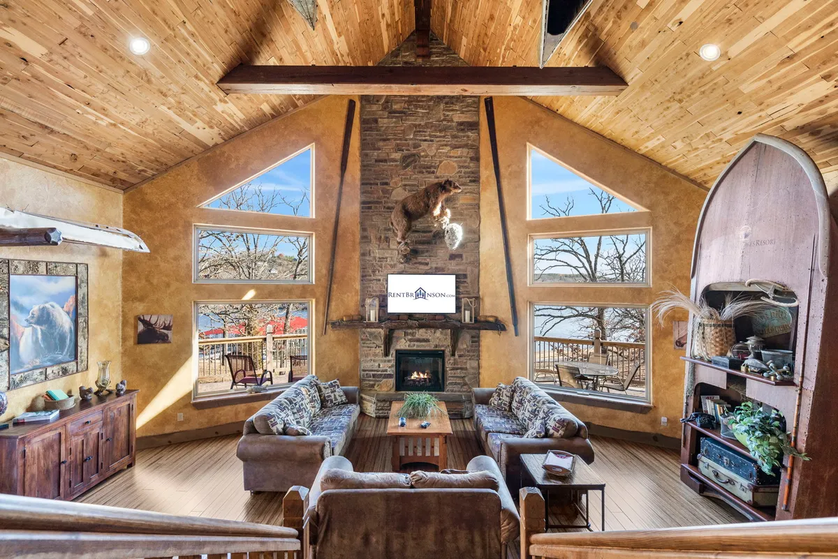 The image showcases a warm, inviting living room centered around a grand stone fireplace, flanked by a majestic taxidermy bear. Expansive windows frame breathtaking lakeside views, while plush seating creates a perfect gathering space for family and friends.