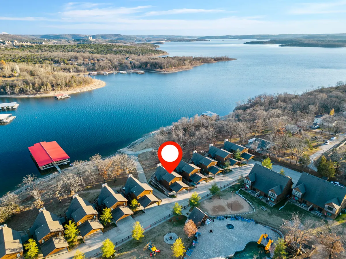 Lakefront cabins line the water with a spacious playground and a boat dock visible in the background. The scene is tranquil, showing the proximity of accommodations to both play and lake access, with room for group activities and lakeside fun.