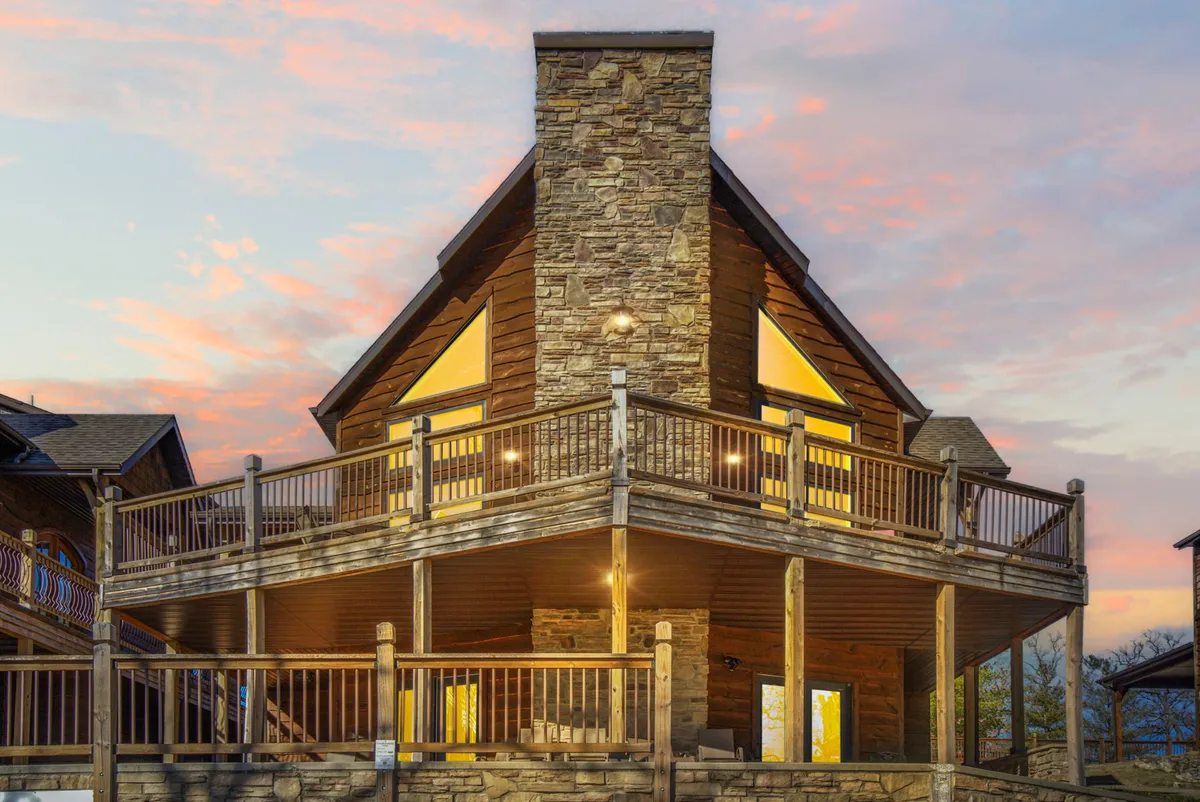 This image showcases an inviting exterior with a stone chimney, large windows, and an expansive wooden deck. The deck is set up for gatherings, offering ample space and seating for groups to dine, socialize, or enjoy serene sunset views over the lake.