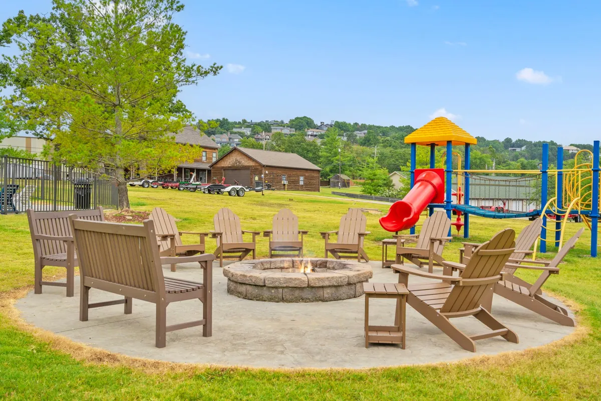 An inviting exterior view of a communal fire pit surrounded by adirondack chairs, set near a playground slide. The cozy setup hints at evening gatherings, storytelling, and casual socializing under the open sky.