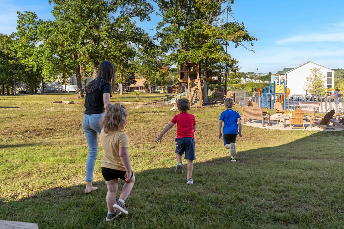 A joyful playground scene where children are climbing and exploring on colorful equipment, with an adult nearby supervising on a well-manicured lawn surrounded by trees. The image conveys laughter, movement, and carefree fun for younger guests.