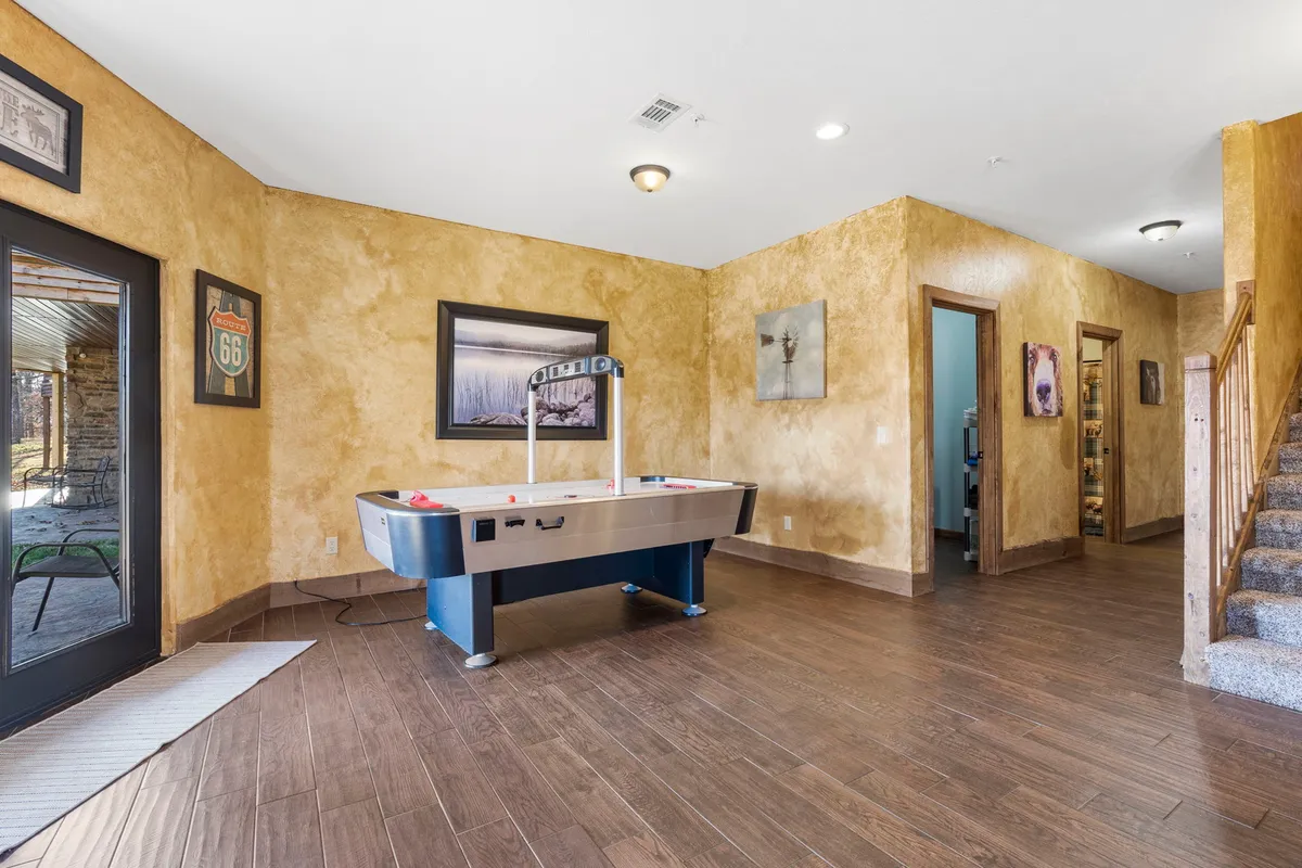 A cheerful and energetic game area with a prominent air hockey table ready for action. Framed wall art accents the space, while a doorway leads out to the patio, hinting at seamless indoor-outdoor entertainment. The staircase in view suggests this is part of a dynamic lower level for group activities.