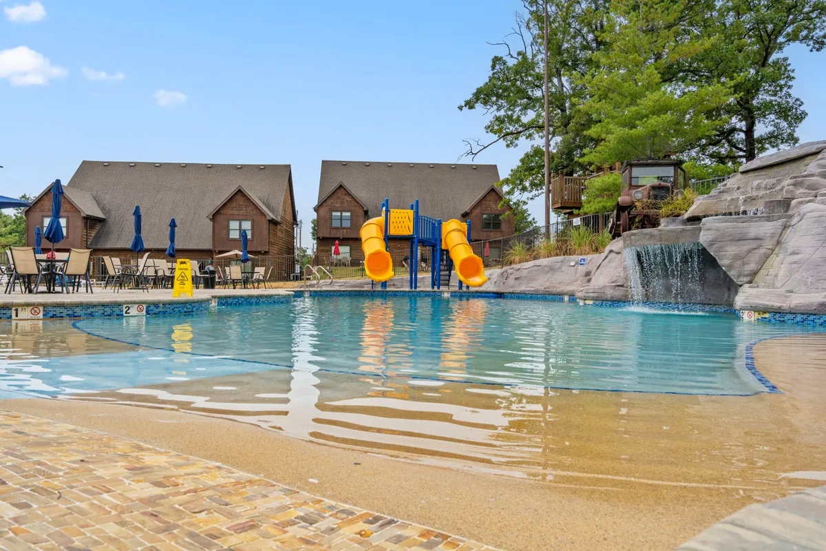 A lively pool scene featuring a large swimming pool with a water slide, a cascading rock waterfall, and nearby cabins. Lounge chairs and playful families make the space feel welcoming and full of activity.