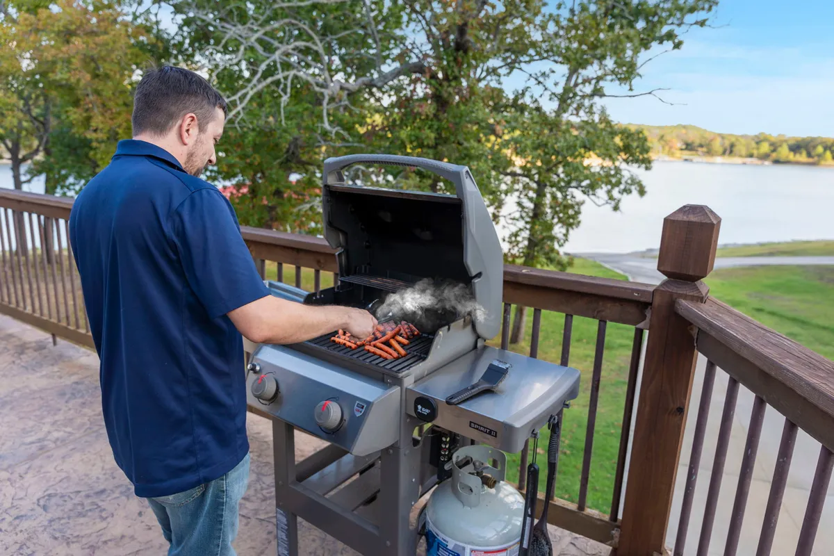 An outdoor deck scene featuring a grill, a person preparing food, a wooden railing, and a sparkling lake in the background. The space radiates casual comfort, suggesting joyful barbecues and shared meals al fresco.