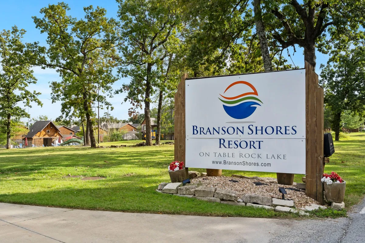 This image presents the Branson Shores Resort sign, surrounded by lush trees and with distant cabins visible in the background. The setting conveys a welcoming resort atmosphere and hints at the proximity to both nature and local attractions.