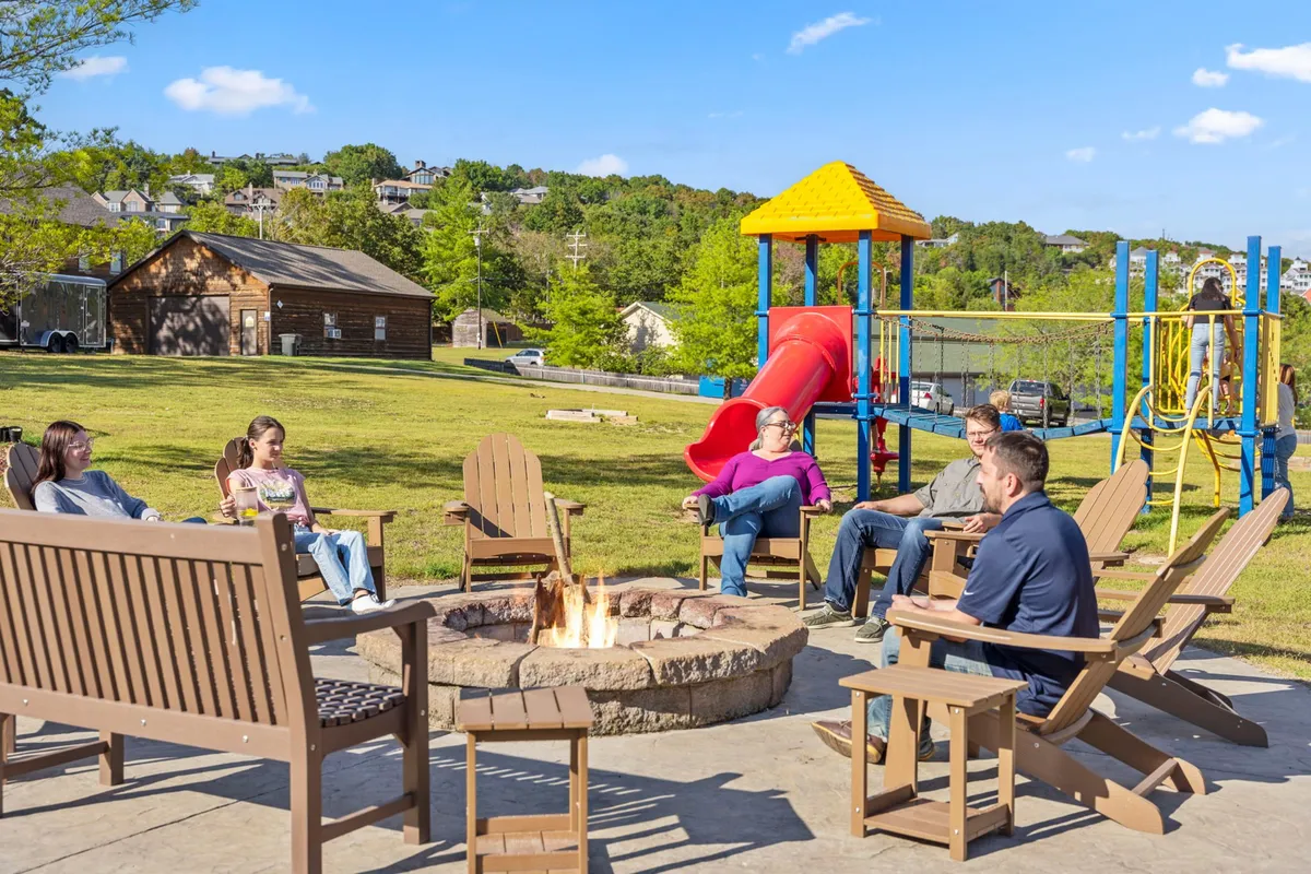 A cheerful gathering around a fire pit near a playground, with adults socializing and children playing freely. The scene shows the warmth and community spirit encouraged by the resort's shared amenities.