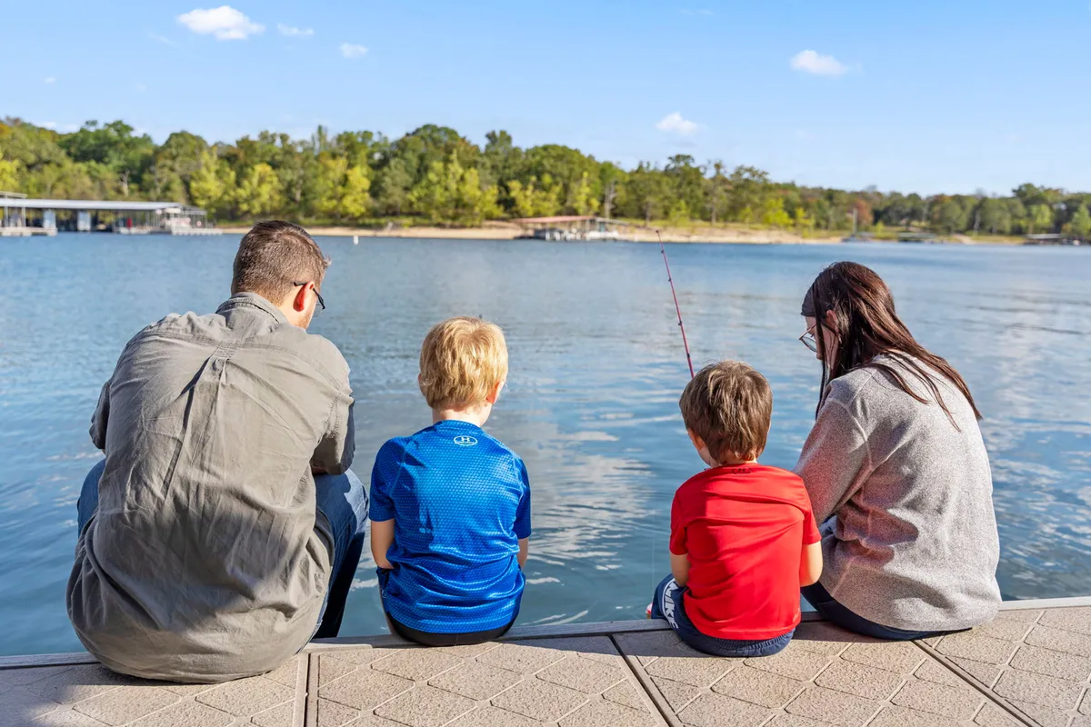 A joyful moment as family members fish together on the calm, tree-fringed lakeshore. The greenery and still water evoke peace and togetherness, while the family's focus on their fishing poles hints at both relaxation and shared adventure.