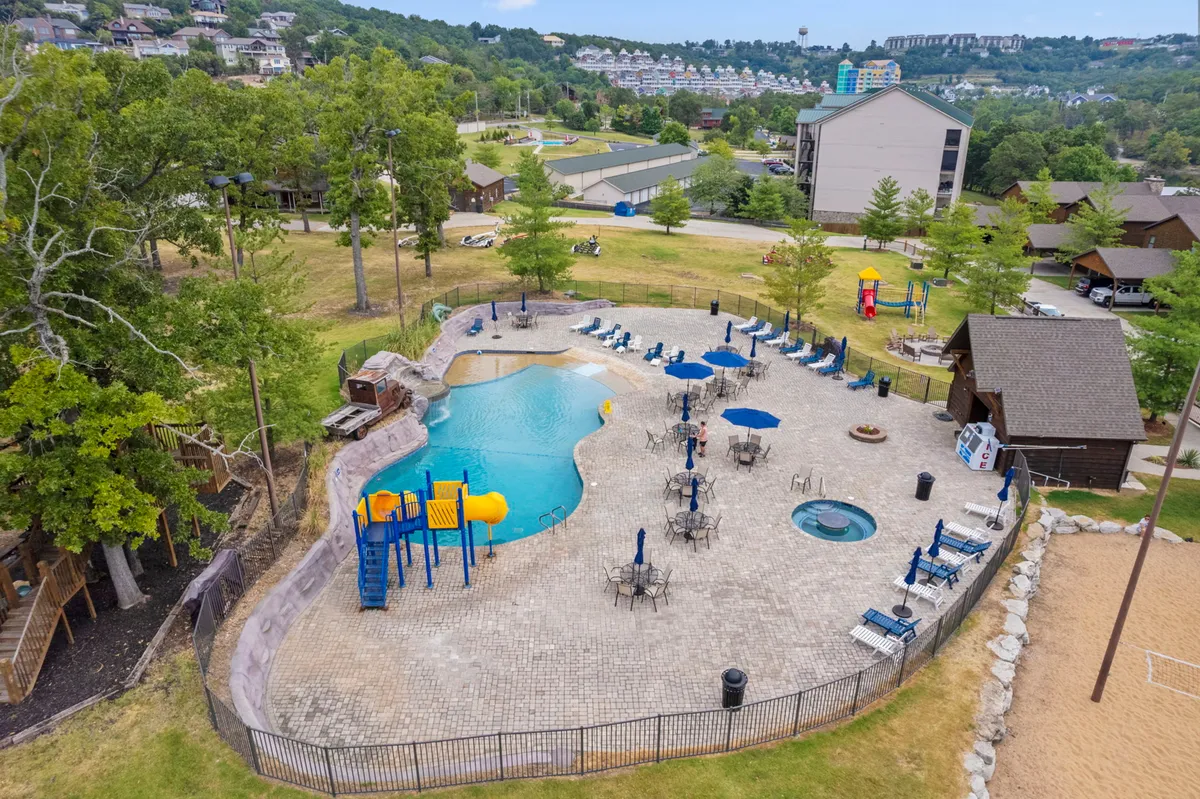 A vibrant outdoor scene featuring a large swimming pool surrounded by lounge chairs and umbrellas, adjacent to a playground with colorful equipment and a hot tub. Children are playing, and families are relaxing in and around the pool area, creating a lively, family-friendly atmosphere.