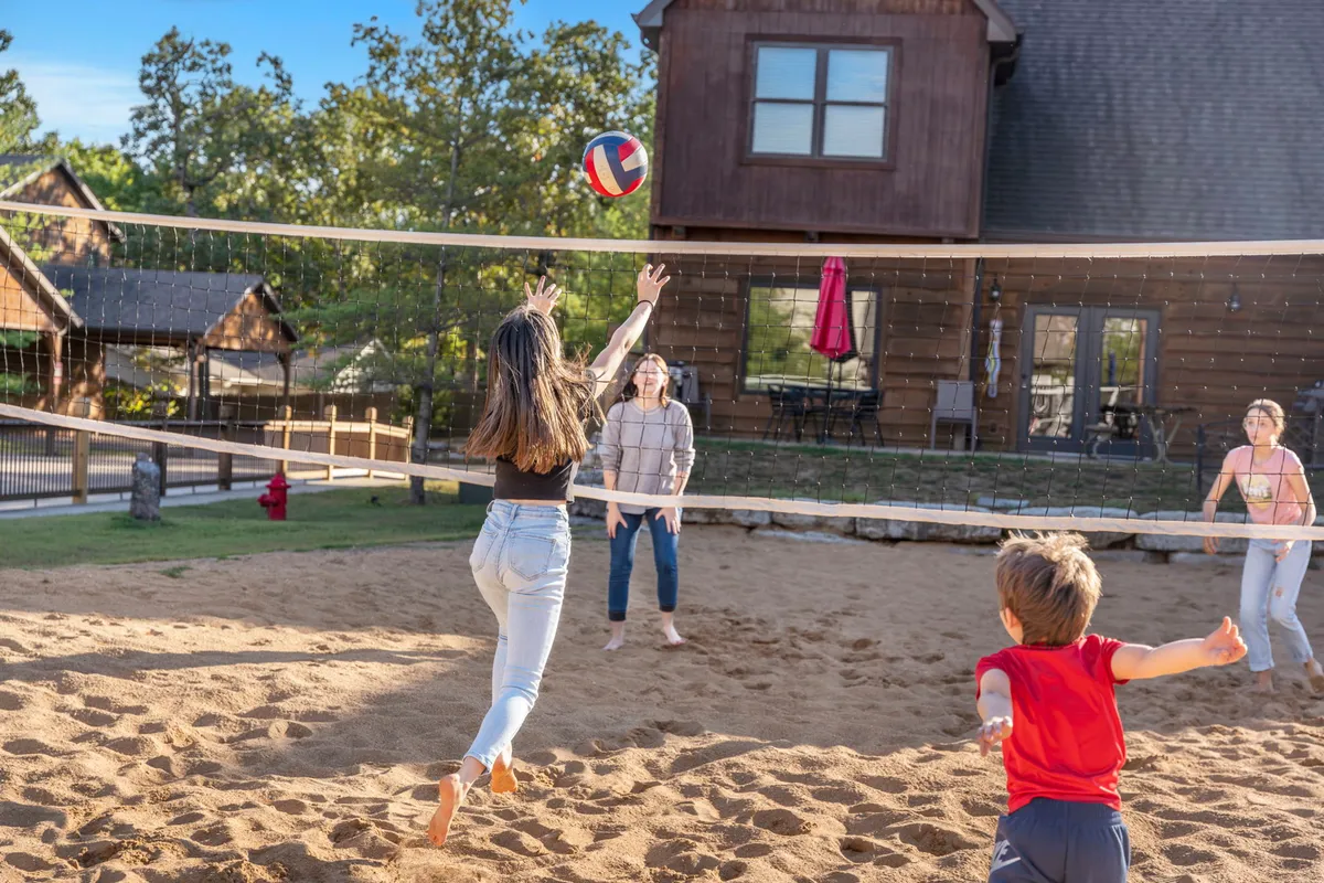 A dynamic view of a sand volleyball court with people actively playing, set against a backdrop of a cozy cabin and sunlit grounds. The image captures movement, camaraderie, and the joy of outdoor sports within the resort.