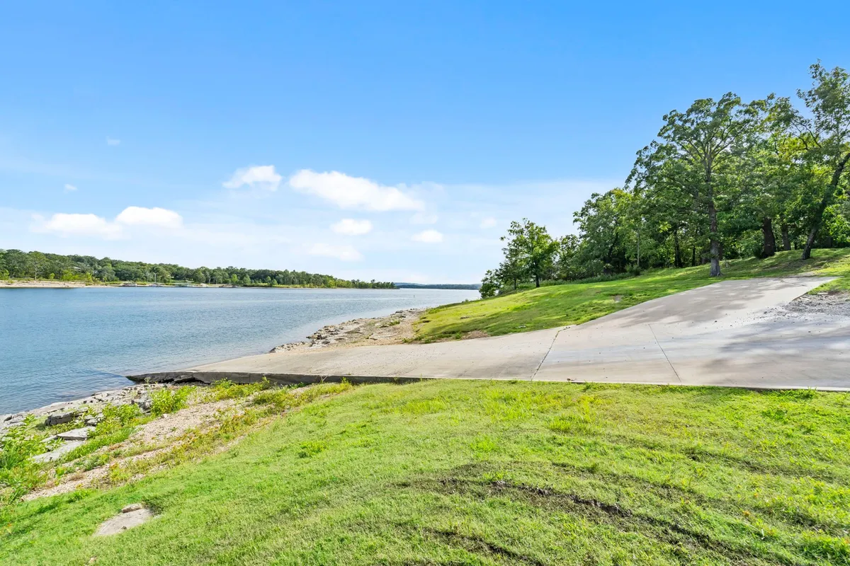 A sweeping waterfront scene with lush green grass leading to the water's edge, bordered by a tree line and featuring a boat ramp. The calm lake reflects the colors of sunset, while the shoreline offers spots for fishing, picnicking, and launching boats or kayaks.