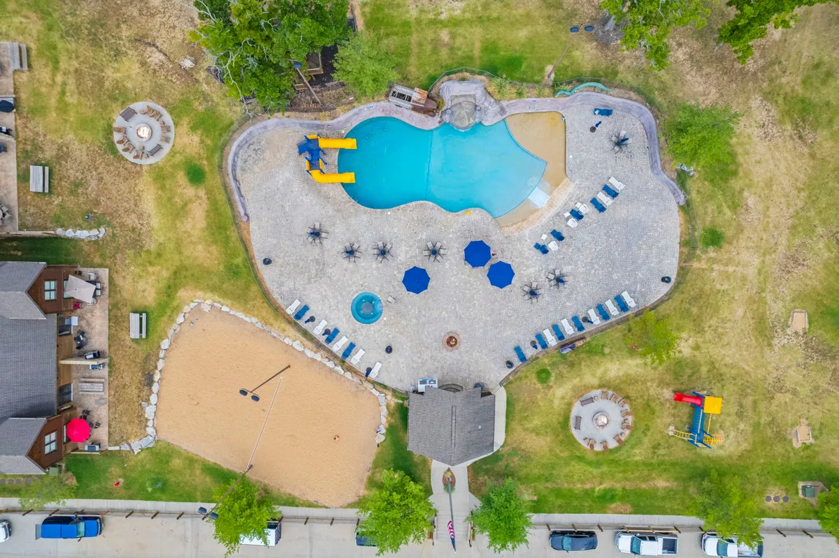 An action-packed exterior view showing a sparkling swimming pool with water slides, a sand volleyball court, a cozy fire pit area, and relaxed seating spaces. The area is bustling with people swimming, playing games, and socializing in a playful and relaxed environment.