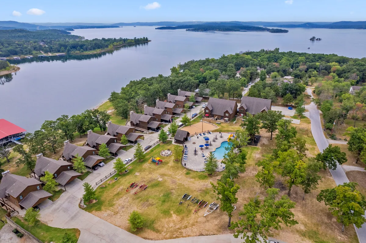 A broad exterior view showing the lake, a swimming pool, multiple cabins, and a playground, all within the same frame. The scene captures families enjoying the pool, children playing, and the tranquil lakeside setting.