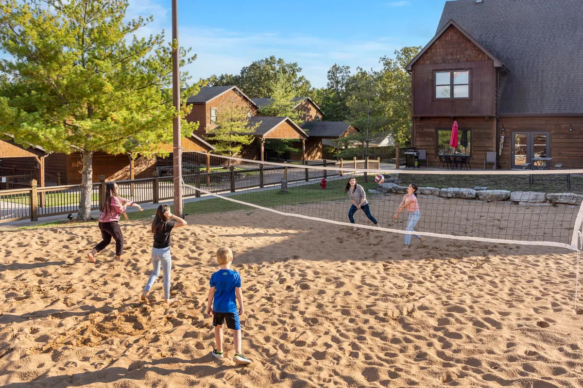 An exterior scene featuring a sand volleyball court surrounded by children playing, family members gathered, and nearby cabins. The area is bathed in sunlight, suggesting a lively and playful atmosphere ideal for all ages.