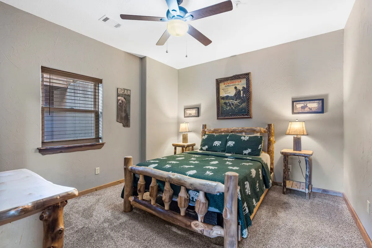 A warmly lit bedroom featuring a rustic log bed with a green patterned blanket, matching nightstands with glowing lamps, and framed nature-themed artwork. A window offers the promise of natural light or stargazing at night, creating a sanctuary-like atmosphere ideal for a master suite.