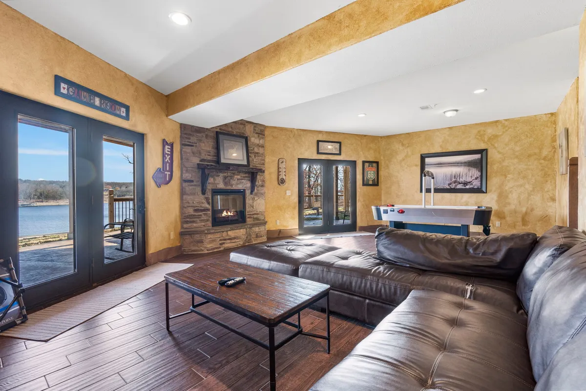 A spacious lower-level living room featuring a large sectional sofa facing a decorative fireplace and a big TV. To the side, a pool table sits ready for friendly competition. The room is bright, with natural light streaming in from glass doors that lead directly outside, offering glimpses of the lake beyond.